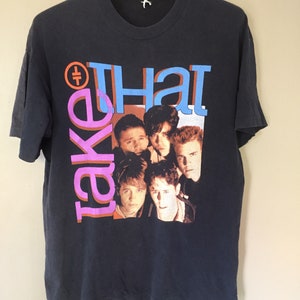 Może przedstawiać: Czarny T-shirt z nadrukiem graficznym zespołu Take That. Członkowie zespołu są przedstawieni na zdjęciu grupowym z nazwą zespołu w dużych, kolorowych literach.