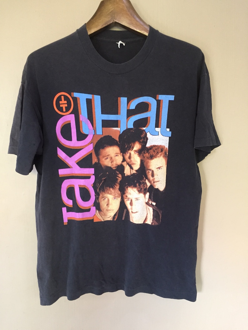 Może przedstawiać: Czarny T-shirt z nadrukiem graficznym zespołu Take That. Członkowie zespołu są przedstawieni na zdjęciu grupowym z nazwą zespołu w dużych, kolorowych literach.