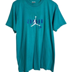 K&ouml;nnte beinhalten: T&uuml;rkisfarbenes T-Shirt mit dem Wort "JORDAN" in blauen Blockbuchstaben und einer wei&szlig;en Silhouette eines Basketballspielers. Ein kleines blaues Nike-Logo befindet sich darunter. Das T-Shirt hat kurze &Auml;rmel und einen Rundhalsausschnitt.