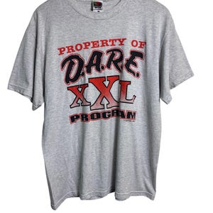 May include: A heather gray t-shirt with the text "PROPERTY OF DARE XXL PROGRAM" in red and black lettering. The shirt is short-sleeved and appears to be made of a soft, comfortable material. The shirt is hanging on a wooden hanger.