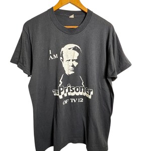 May include: Dark gray t-shirt with a white graphic of a man's face and the text "I AM THE PRISONER OF TV 12." The shirt is hanging on a wooden hanger.