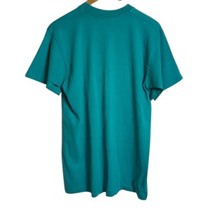 K&ouml;nnte beinhalten: Ein t&uuml;rkisfarbenes T-Shirt h&auml;ngt an einem Holzkleiderb&uuml;gel vor wei&szlig;em Hintergrund. Das kurz&auml;rmelige Shirt ist aus einem weichen Material gefertigt. Das T-Shirt ist einfarbig und scheint ungetragen zu sein.