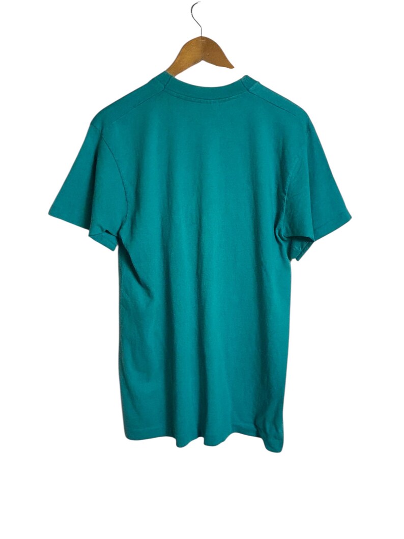 K&ouml;nnte beinhalten: Ein t&uuml;rkisfarbenes T-Shirt h&auml;ngt an einem Holzkleiderb&uuml;gel vor wei&szlig;em Hintergrund. Das kurz&auml;rmelige Shirt ist aus einem weichen Material gefertigt. Das T-Shirt ist einfarbig und scheint ungetragen zu sein.