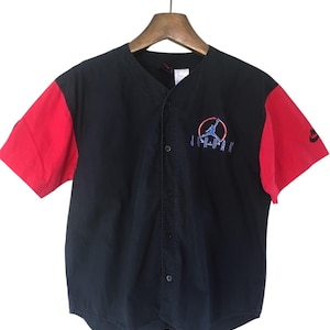 Puede incluir: Una camiseta de b&eacute;isbol negra y roja con un logotipo "JORDAN" blanco y una silueta de jugador de baloncesto en un c&iacute;rculo. Las mangas son rojas y el cuerpo de la camiseta es negro.