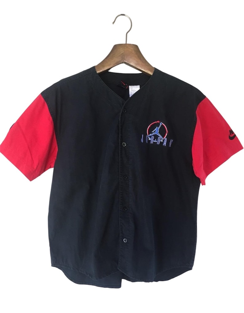 Puede incluir: Una camiseta de b&eacute;isbol negra y roja con un logotipo "JORDAN" blanco y una silueta de jugador de baloncesto en un c&iacute;rculo. Las mangas son rojas y el cuerpo de la camiseta es negro.