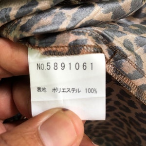 May include: A white tag with black text that reads "NO. 5891061" and "表地 ポリエステル 100%". The tag is attached to a brown and black leopard print fabric.
