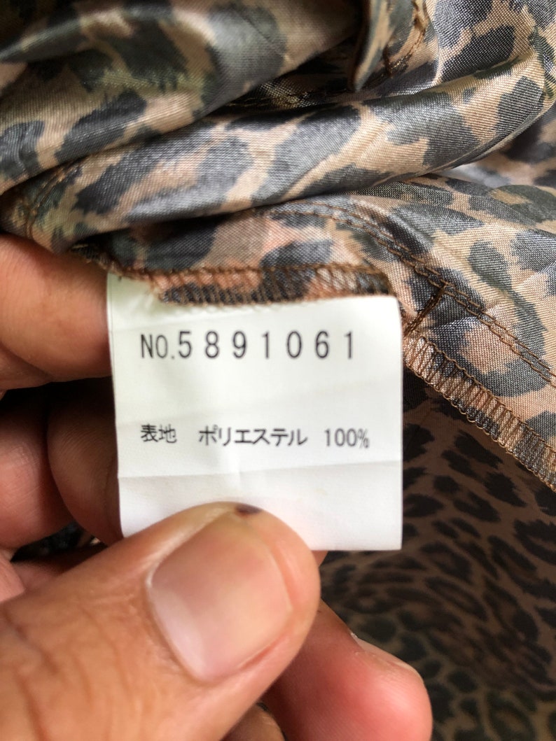 May include: A white tag with black text that reads "NO. 5891061" and "表地 ポリエステル 100%". The tag is attached to a brown and black leopard print fabric.