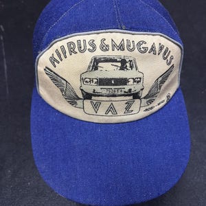 Puede incluir: Una gorra de béisbol azul con un parche beige que presenta una ilustración en blanco y negro de un automóvil con alas y el texto "KIPRUS & MUGAVUS VAZ ROBIN-senor".