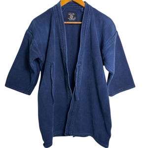 Peut inclure: Veste de style kimono bleu marine avec des manches trois-quarts suspendue à un cintre en bois. La veste a un tissage texturé et une fermeture à nouer. Une petite étiquette est visible près du col.