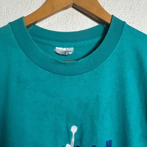 K&ouml;nnte beinhalten: T&uuml;rkisfarbenes Sweatshirt mit Rundhalsausschnitt, auf dem das Wort "JORDAN" in Blau und eine wei&szlig;e Silhouette eines Basketballspielers gestickt ist. Das Shirt h&auml;ngt an einem Holzkleiderb&uuml;gel.