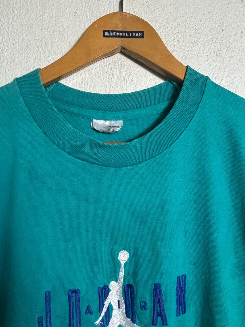 K&ouml;nnte beinhalten: T&uuml;rkisfarbenes Sweatshirt mit Rundhalsausschnitt, auf dem das Wort "JORDAN" in Blau und eine wei&szlig;e Silhouette eines Basketballspielers gestickt ist. Das Shirt h&auml;ngt an einem Holzkleiderb&uuml;gel.