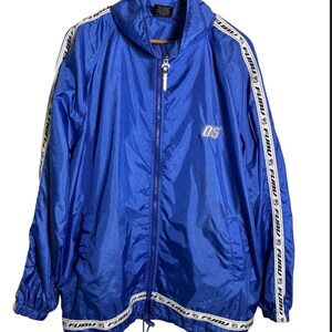 Op de afbeelding: Een felblauw windjack met een volledige ritssluiting aan de voorzijde, een hoge kraag en lange mouwen. Witte strepen lopen langs de mouwen en de onderzoom, bedrukt met de herhalende zwarte tekst FUBU. Het getal 05 is in het wit op de linkerborst geborduurd.