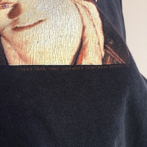 Może przedstawiać: Czarny T-shirt z wyblakłym obrazem twarzy mężczyzny. Na dole koszulki nadrukowany jest tekst "1999 Take That Licensed To Universal Music Ltd."