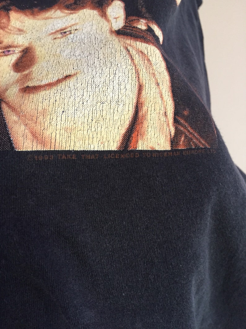 Może przedstawiać: Czarny T-shirt z wyblakłym obrazem twarzy mężczyzny. Na dole koszulki nadrukowany jest tekst "1999 Take That Licensed To Universal Music Ltd."