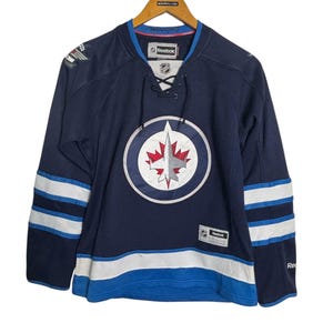 May include: Navy blue hockey jersey with white and light blue stripes on the sleeves and bottom. The jersey features a circular logo with a maple leaf and the text "Winnipeg" on the collar. The Reebok logo is visible.