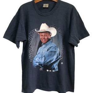 May include: A vintage navy blue t-shirt featuring a printed image of a man wearing a cowboy hat and a blue denim shirt. The man is smiling and has his arms crossed. The background is a faded, distressed pattern.