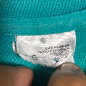 K&ouml;nnte beinhalten: Nahaufnahme eines t&uuml;rkisfarbenen T-Shirt-Kragens mit einem wei&szlig;en Etikett. Auf dem Etikett steht "MADE IN CANADA" und "FABRIQUE AU CANADA" mit "100% COTTON" darunter. Der Stoff des Shirts hat eine gerippte Textur.