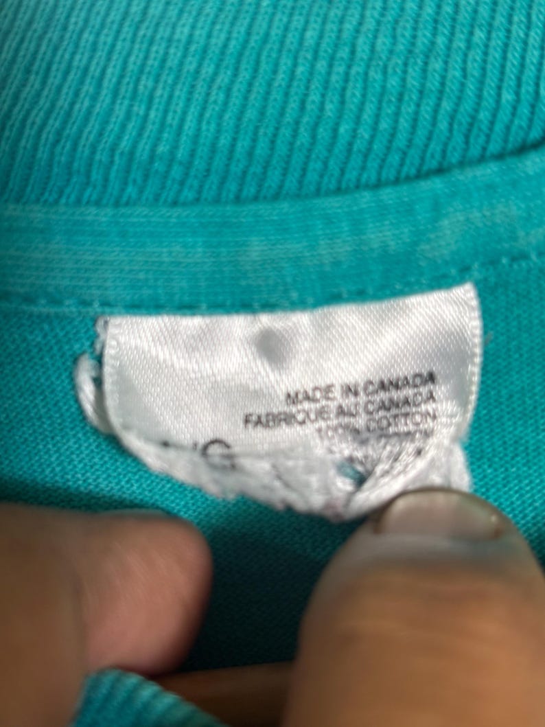 K&ouml;nnte beinhalten: Nahaufnahme eines t&uuml;rkisfarbenen T-Shirt-Kragens mit einem wei&szlig;en Etikett. Auf dem Etikett steht "MADE IN CANADA" und "FABRIQUE AU CANADA" mit "100% COTTON" darunter. Der Stoff des Shirts hat eine gerippte Textur.