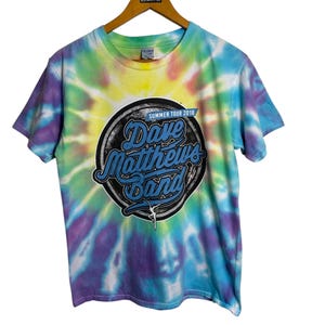 Può includere: Una t-shirt tie-dye con un logo circolare che presenta il testo "Dave Matthews Band" e "Summer Tour 2018". Il logo è in blu, nero e bianco su sfondo nero.