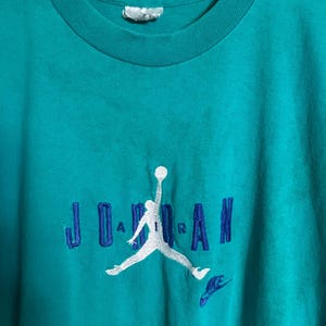 K&ouml;nnte beinhalten: T&uuml;rkisfarbenes T-Shirt mit dem Wort "JORDAN" in Blau gestickt, zusammen mit einer wei&szlig;en Silhouette eines Basketballspielers und einem kleinen blauen Nike-Logo. Das Shirt hat einen Rundhalsausschnitt und kurze &Auml;rmel.