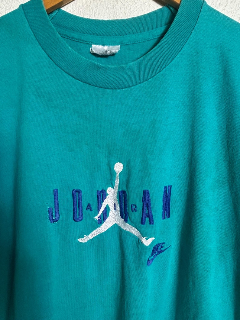 K&ouml;nnte beinhalten: T&uuml;rkisfarbenes T-Shirt mit dem Wort "JORDAN" in Blau gestickt, zusammen mit einer wei&szlig;en Silhouette eines Basketballspielers und einem kleinen blauen Nike-Logo. Das Shirt hat einen Rundhalsausschnitt und kurze &Auml;rmel.