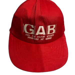 May include: A vibrant red baseball cap with a curved peak. The cap displays the white embroidered text "GAB" above the words "FEEL THE MOTOR SPORTS PRODUCED BY WINGS". The cap is made of a smooth fabric and has a rounded crown.