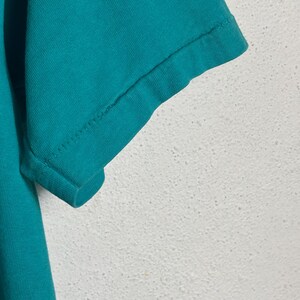 K&ouml;nnte beinhalten: Nahaufnahme eines t&uuml;rkisfarbenen T-Shirt-&Auml;rmels. Der &Auml;rmel ist am Saum umgeschlagen, wobei die N&auml;hte zu sehen sind. Der Stoff scheint ein weiches Baumwollmaterial zu sein. Der Hintergrund ist eine strukturierte wei&szlig;e Oberfl&auml;che.