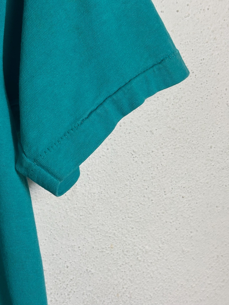 K&ouml;nnte beinhalten: Nahaufnahme eines t&uuml;rkisfarbenen T-Shirt-&Auml;rmels. Der &Auml;rmel ist am Saum umgeschlagen, wobei die N&auml;hte zu sehen sind. Der Stoff scheint ein weiches Baumwollmaterial zu sein. Der Hintergrund ist eine strukturierte wei&szlig;e Oberfl&auml;che.