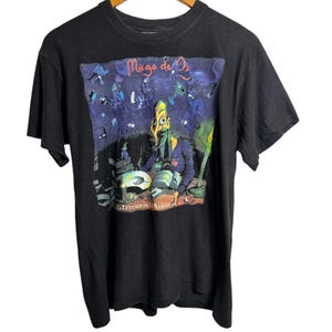 May include: Black t-shirt featuring a colorful graphic from "Mago de Oz." The design includes a cartoonish figure with a long nose and a purple background with various creatures. The text "LA LEYENDA DE LA MANCHA" is also visible.