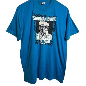 May include: A turquoise blue t-shirt featuring a graphic of Sherman Evans & Band. The graphic is a black and white portrait of a man wearing a hat and sunglasses, with the band's name in bold white letters. The shirt is hanging on a wooden hanger.