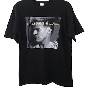 May include: Black t-shirt with a black and white photo of Bryan Adams on the front. The text "Bryan Adams Bare Bones" is printed in gold on the photo.