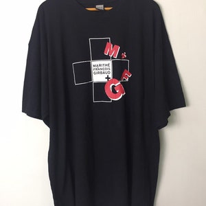 May include: Black t-shirt with a white cross graphic and red text that reads "MARITHÉ + FRANÇOIS GIRBAUD" and "M+G" in a bold, stylized font.