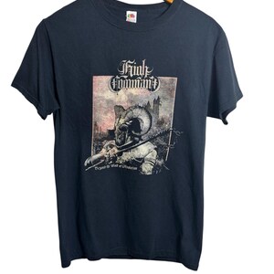 May include: Black t-shirt with a graphic featuring a horned figure holding a weapon, set against a castle backdrop. The text "Trial Command" is above the image, with "Beyond the Wall of Protection" below. The shirt is hanging on a wooden hanger.