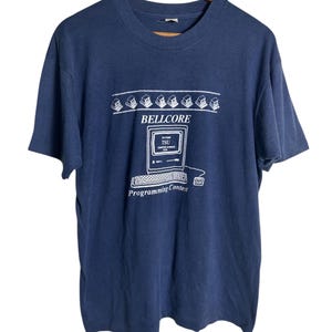 Peut inclure: T-shirt bleu marine avec un motif blanc représentant un ordinateur et le texte "BELLCORE Programming Contest". Le motif comprend une rangée d'icônes d'ordinateurs au-dessus du texte. Le t-shirt est suspendu à un cintre en bois.