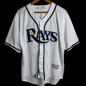 May include: White baseball jersey with the word "RAYS" in navy blue and light blue across the chest. The jersey has navy blue trim and a star logo. The jersey is button-down and has short sleeves.