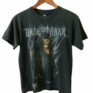 May include: A black t-shirt with a graphic print of The Undertaker, a professional wrestler, wearing a black leather jacket and hat. The text "UNDERTAKER" is printed in a gothic font above the image.