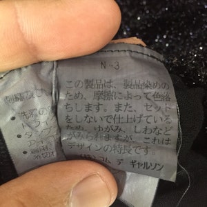 May include: A black and white tag with the text "N-3" and a warning about color fading due to friction. The tag also mentions that the garment is intentionally designed with wrinkles and creases. The tag is labeled "(株)コムデギャルソン", which is the Japanese name for Comme des Gar&ccedil;ons.
