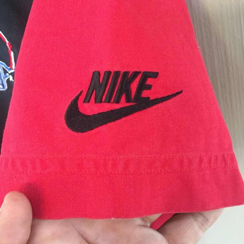 Puede incluir: Camiseta roja con un logotipo de Nike negro y la palabra "NIKE" bordada en el lado derecho de la camiseta.