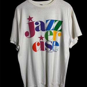 May include: A vintage off-white t-shirt with the words "Jazzercise" in colorful, stylized lettering. The design includes stars and a rainbow of colors, with the brand name "Budi Shep Missett" visible.
