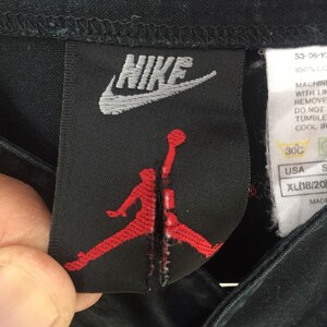 Puede incluir: Camiseta Nike negra con un logotipo Jumpman rojo y blanco. La etiqueta dice "NIKE" e incluye instrucciones de cuidado.