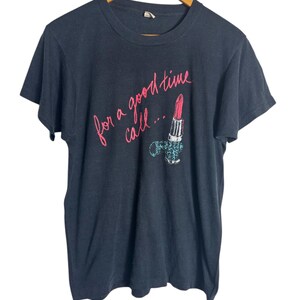 Può includere: T-shirt nero con la scritta "for a good time call..." in corsivo rosa. La maglietta presenta una grafica di un rossetto e di un oggetto decorativo. La maglietta è appesa a una gruccia di legno.