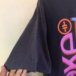 Może przedstawiać: Szary t-shirt z kolorowym nadrukiem graficznym z tyłu. Grafika przedstawia r&oacute;żowy, pomarańczowy i niebieski wz&oacute;r z tekstem "The" i symbolem.