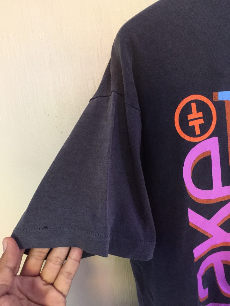 Może przedstawiać: Szary t-shirt z kolorowym nadrukiem graficznym z tyłu. Grafika przedstawia r&oacute;żowy, pomarańczowy i niebieski wz&oacute;r z tekstem "The" i symbolem.