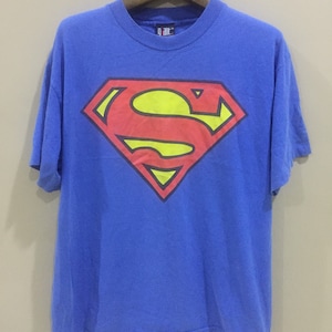 以下が含まれることがあります： 前面に赤と黄色のスーパーマンのロゴがプリントされた青いTシャツです。