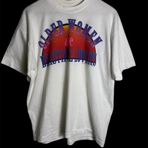 May include: White t-shirt with the text "OLDER WOMEN are BEAUTIFUL LOVERS" in a colorful, arched design. The text is in purple and blue, with a sunset-like gradient background. The shirt is hanging on a wooden hanger.