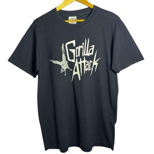 May include: Black t-shirt with the text "Gorilla Attack" in a stylized font, accompanied by a graphic of a pterodactyl-like creature. The shirt is hanging on a wooden hanger.