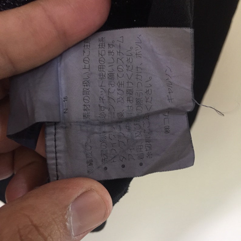 May include: A black and white care tag with instructions for washing and dry cleaning a garment. The tag includes the text "N-16", "刺絡及び", "素材の取扱い上のご注", "必ずネット使用の石油系", "ハライクリーニングでお願いします。", "・タンブラー、及び全てのスチーム", "アイコンはお避けください。", "●着用導及び所の際引っかけ、ホツレ、", "糸切根にこください。", "(株)コム", "ギャルソン".