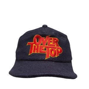 Könnte beinhalten: Marineblaue Kappe mit dem gestickten Text "OVER THE TOP" in Rot und Gelb. Der Hut hat eine runde Krone und einen kurzen Schirm. Der Text ist in einer fetten, stilisierten Schrift.