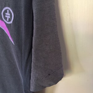 Może przedstawiać: Szary t-shirt z nadrukiem w kolorze r&oacute;żowym i białym przedstawiającym symbol z krzyżem w k&oacute;łku. T-shirt ma kr&oacute;tki rękaw i żebrowany kołnierzyk.
