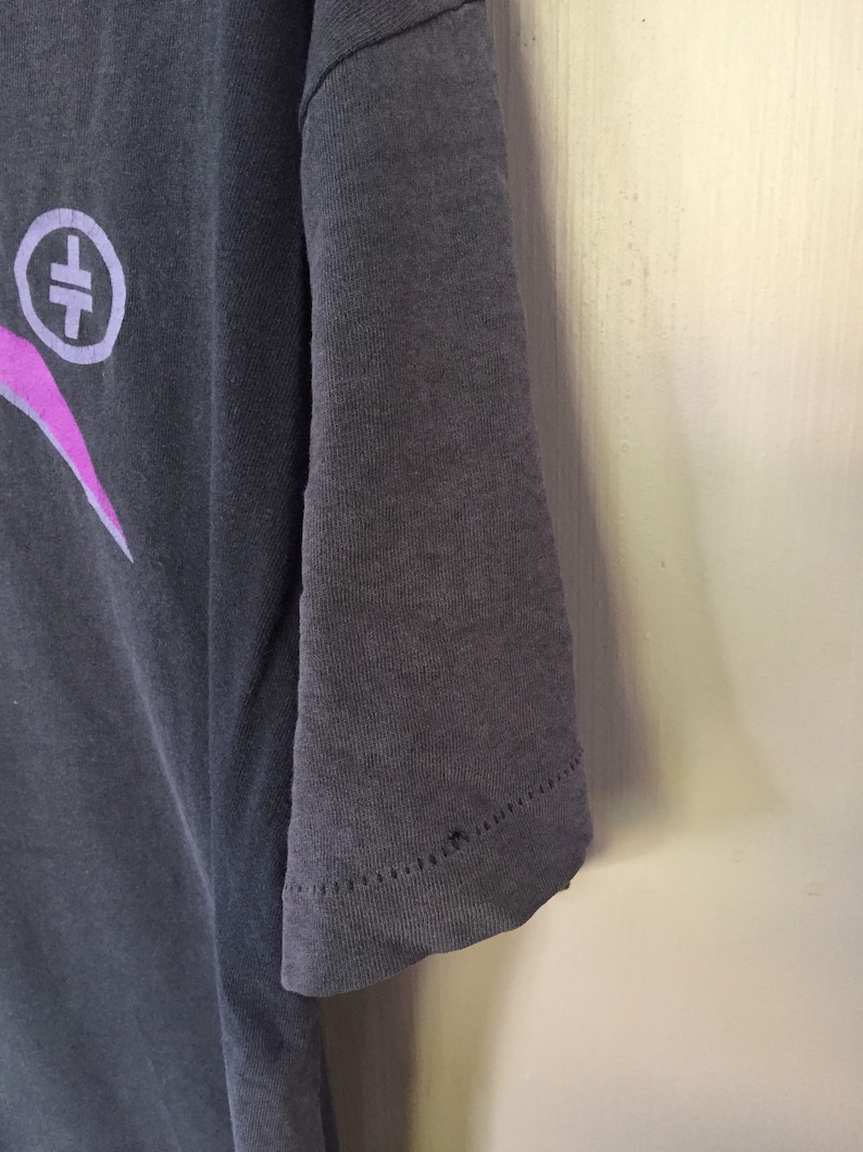 Może przedstawiać: Szary t-shirt z nadrukiem w kolorze r&oacute;żowym i białym przedstawiającym symbol z krzyżem w k&oacute;łku. T-shirt ma kr&oacute;tki rękaw i żebrowany kołnierzyk.
